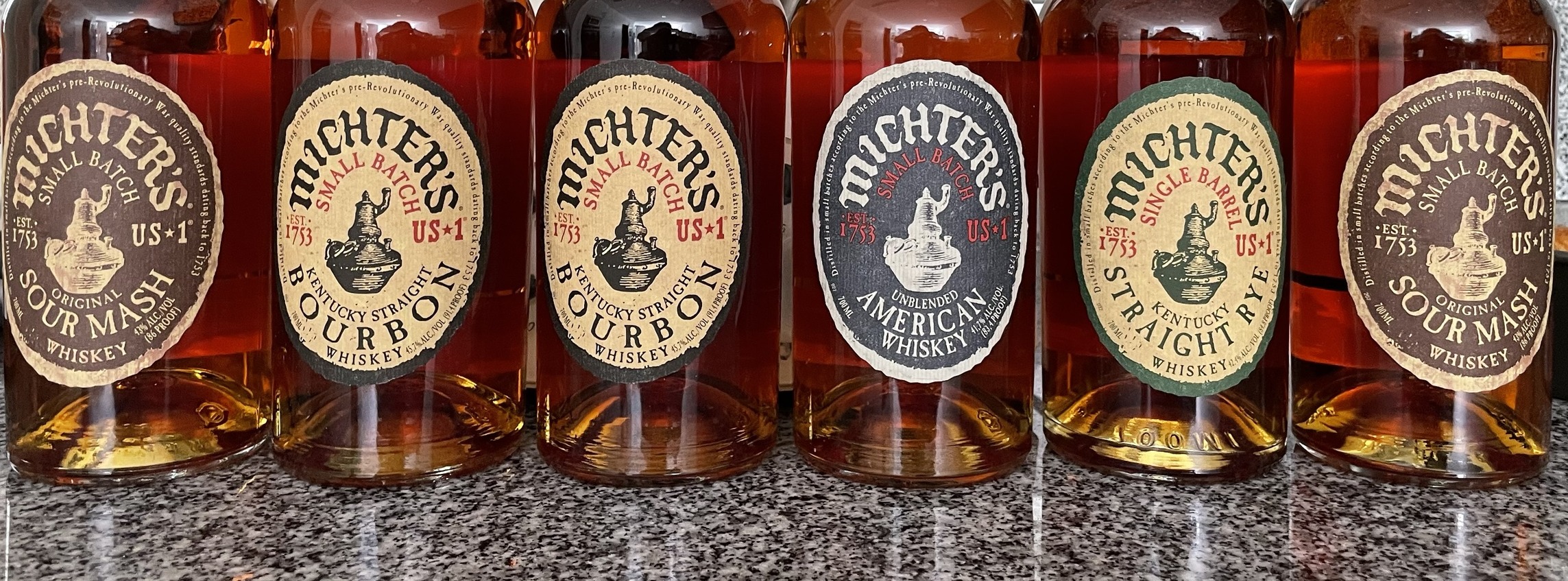 Seis garrafas de Michter’s US★1 alinhadas numa bancada de granito, com os rótulos à vista